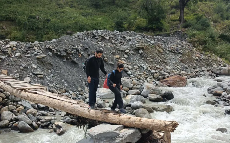 Hikers crossing a rushing mountain stream over fallen tree logs on the Beas Kund trail