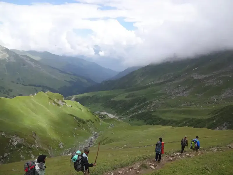 Trekkers hiking across vast alpine meadows with misty clouds over Bara Bhangal valley