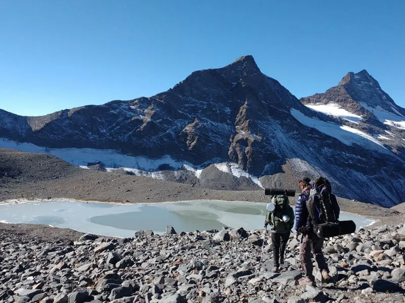 Two trekkers carrying backpacks on a high-altitude rocky moraine near a glacial lake