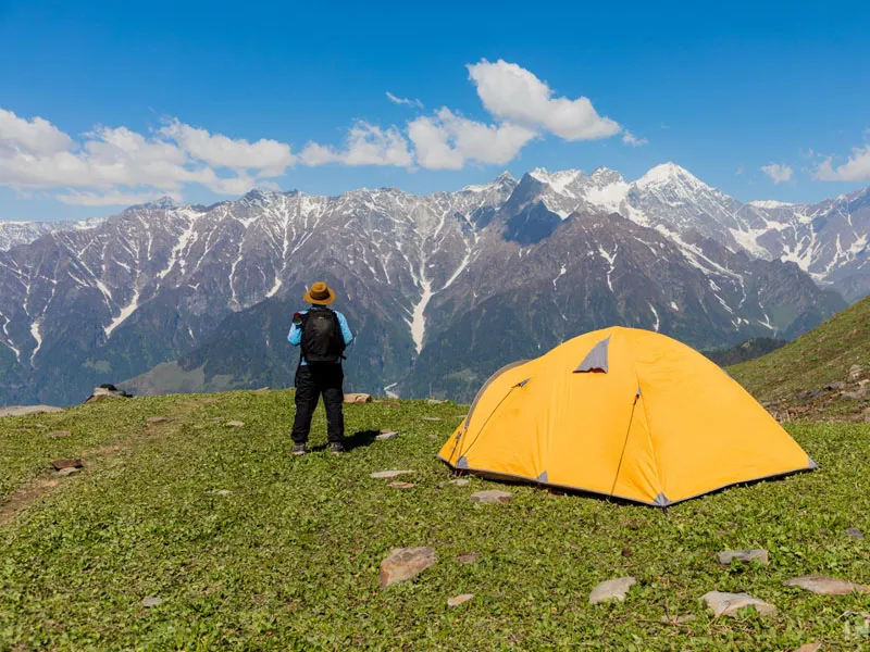 Solo trekker standing beside a yellow alpine tent facing snow-capped Himalayan ranges
