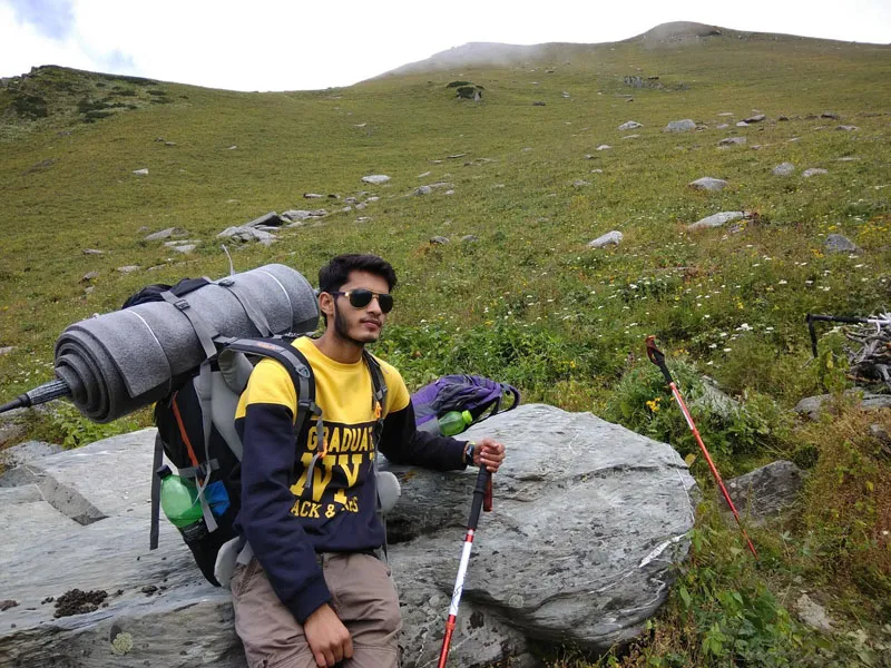 Trekker with full backpack and trekking poles resting on a rock along the Dehnasar Lake trail
