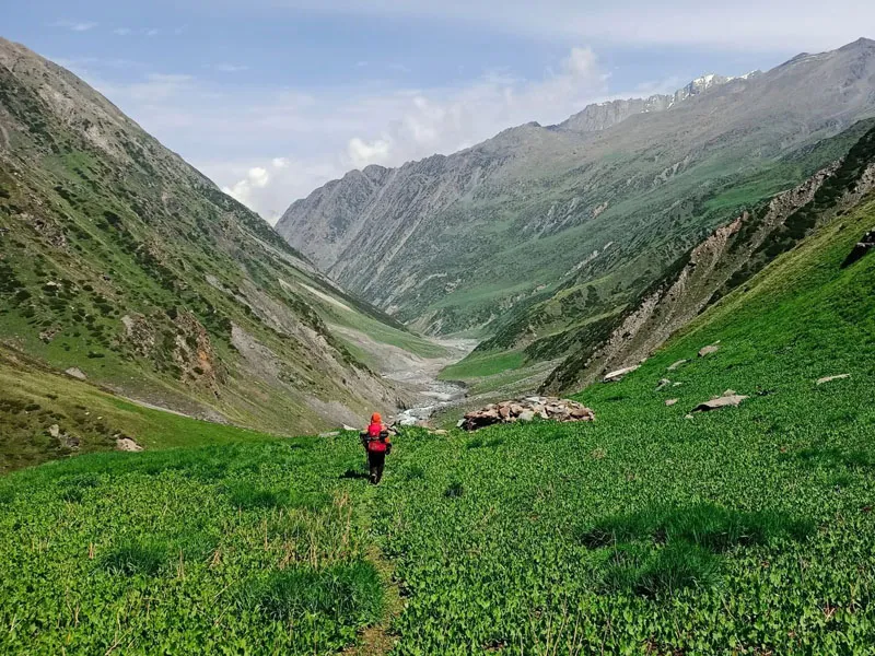 Solo trekker walking along a vast lush green mountain valley flanked by towering slopes