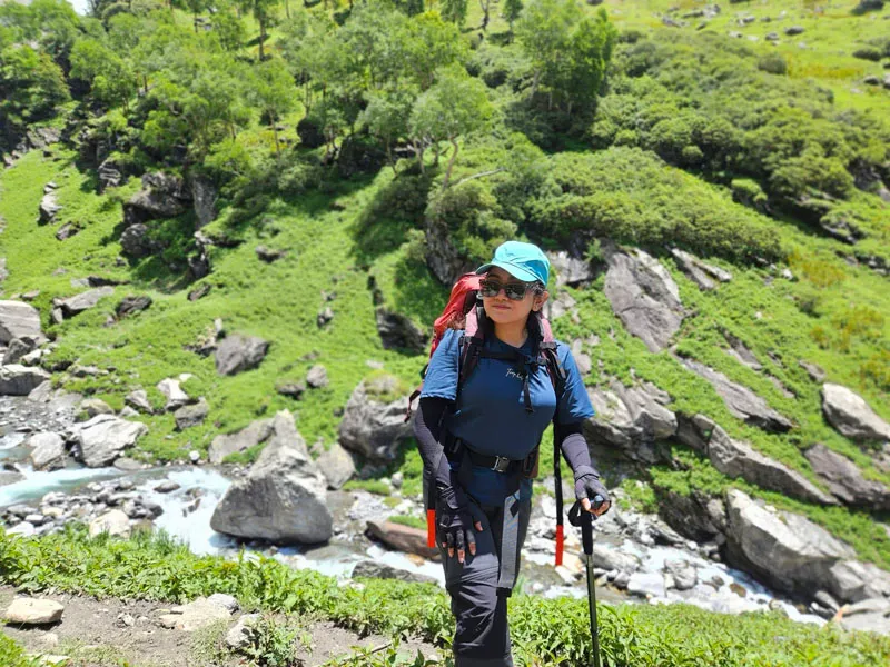 Female trekker with heavy backpack and trekking pole smiling beside a mountain stream in Balu Ka Ghera