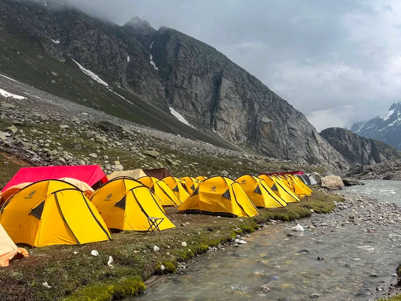 Line of bright yellow alpine tents pitched along a stream beneath steep rocky mountains at Shea Goru