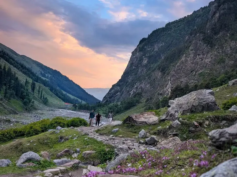 Hikers walking along a rocky valley path beneath a glowing sunset sky in the Himalayas