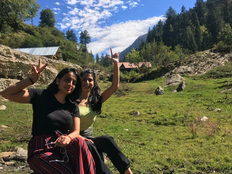 Two girls posing happily on green meadows surrounded by tall pine trees in Kheerganga