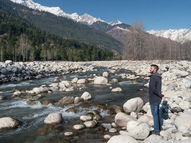 Traveler standing on the rocky banks of the Beas River facing snow-capped Himalayan mountain peaks