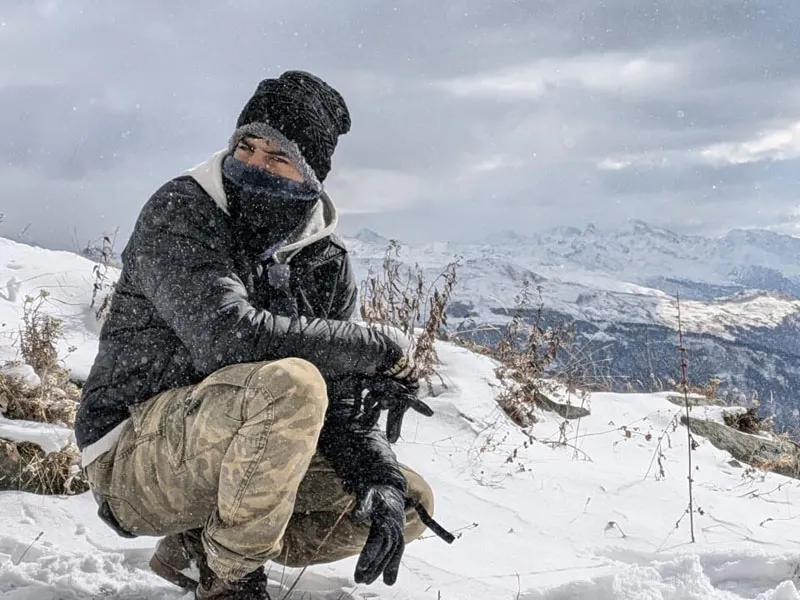 Trekker bundled up in warm winter gear crouching on snow-covered ridge on Patalsu Peak trek