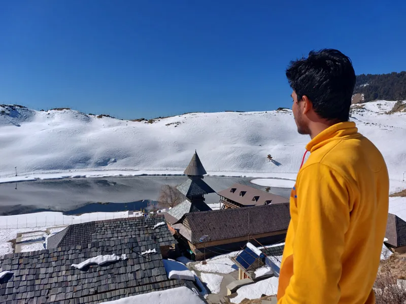 Trekker in yellow jacket looking out over snow-covered Prashar Lake basin and ancient pagoda temple