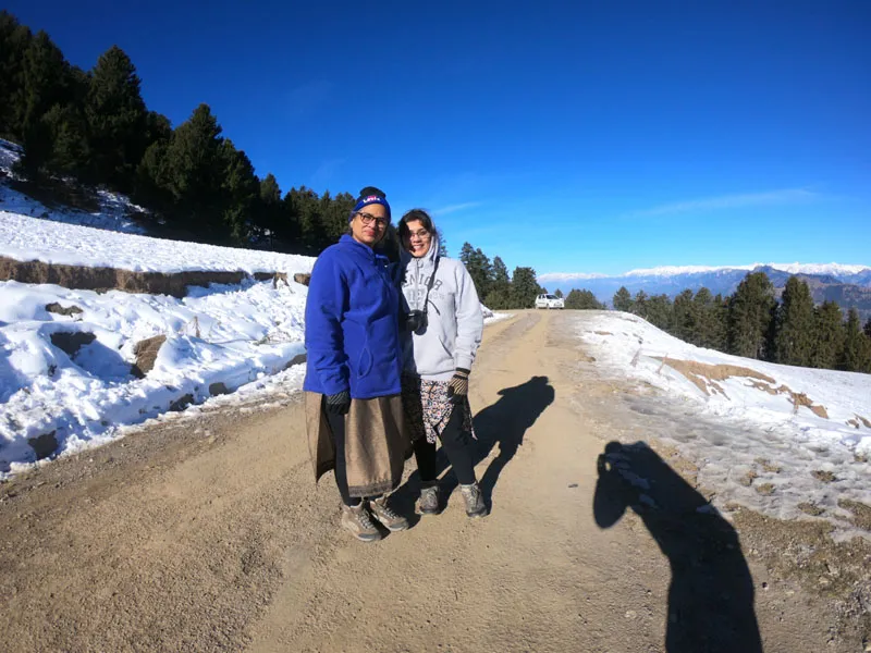 Two travelers standing on a high mountain road flanked by snow patches and pine trees under a clear blue sky