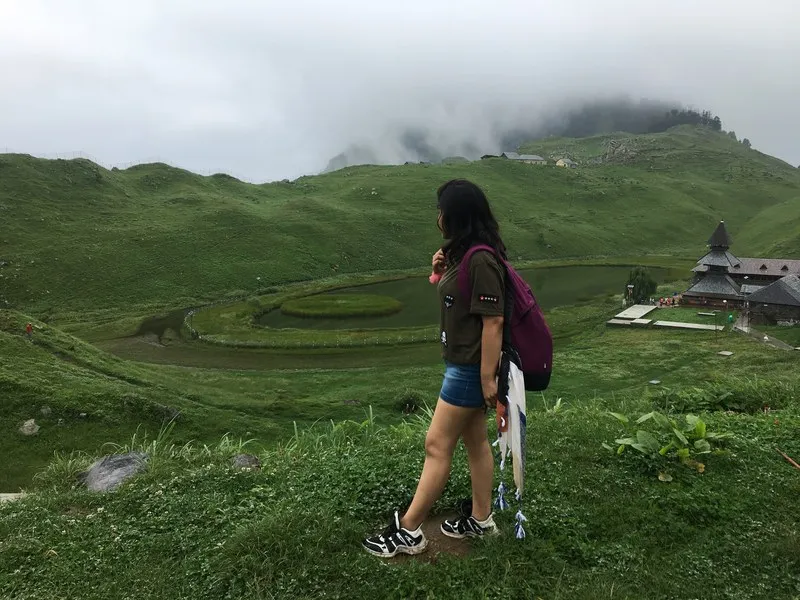 Solo female trekker standing on lush green rolling meadows overlooking Prashar Lake valley