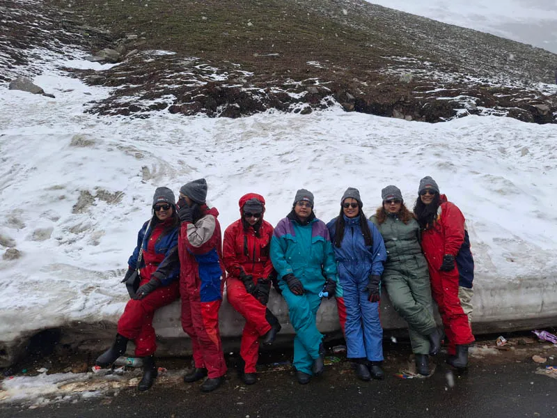 Tourists in colorful snowsuits sitting beside a tall snow wall at Rohtang Pass near Manali