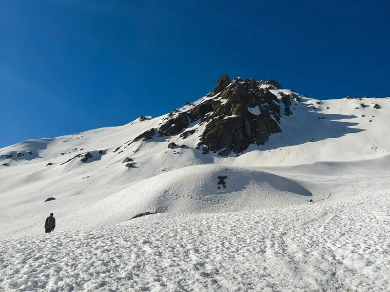 Trekkers ascending a massive snow-covered slope beneath sharp rocky mountain peaks