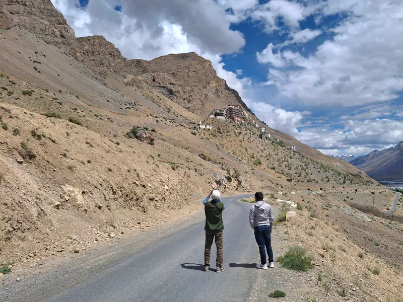 Two travelers standing on an asphalt road winding through the rugged arid terrain of Spiti Valley