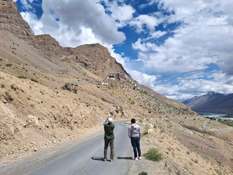 Two travelers standing on an empty asphalt mountain road winding through the cold desert landscape of Spiti