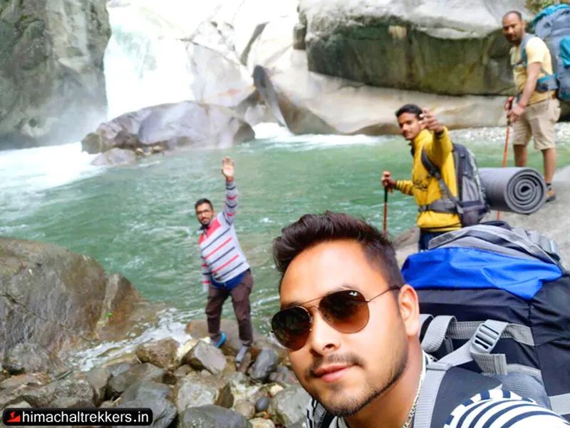 Trekkers taking a selfie beside a pristine turquoise river pool and waterfall in Tirthan Valley