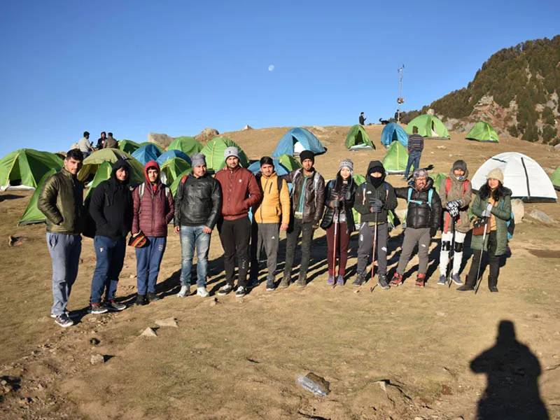 Large trekking group standing in line at Triund hilltop campsite dotted with green tents