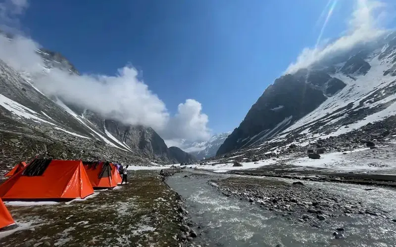 Orange campsite tents pitched near a glacial river bed nestled between steep rocky mountains at Hampta Pass