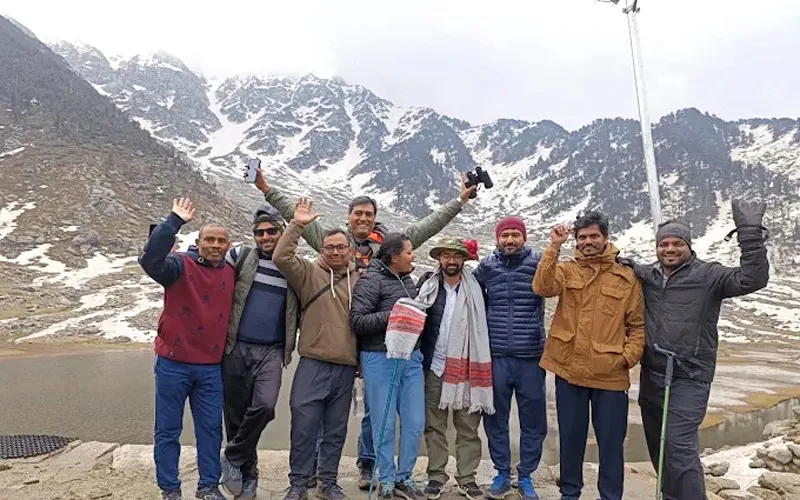 Group of trekkers raising hands joyfully against the snow-dusted Dhauladhar mountain range at Kareri Lake