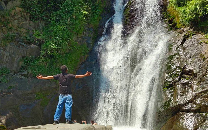 Trekker with arms raised facing the powerful rushing stream of Lapas Waterfall in Barot Valley