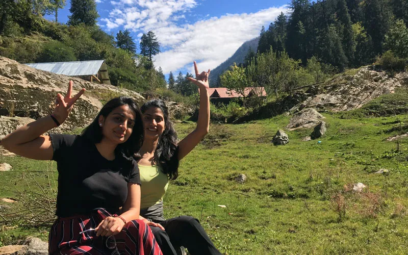 Two female hikers relaxing and gesturing happily on green meadows framed by pine trees