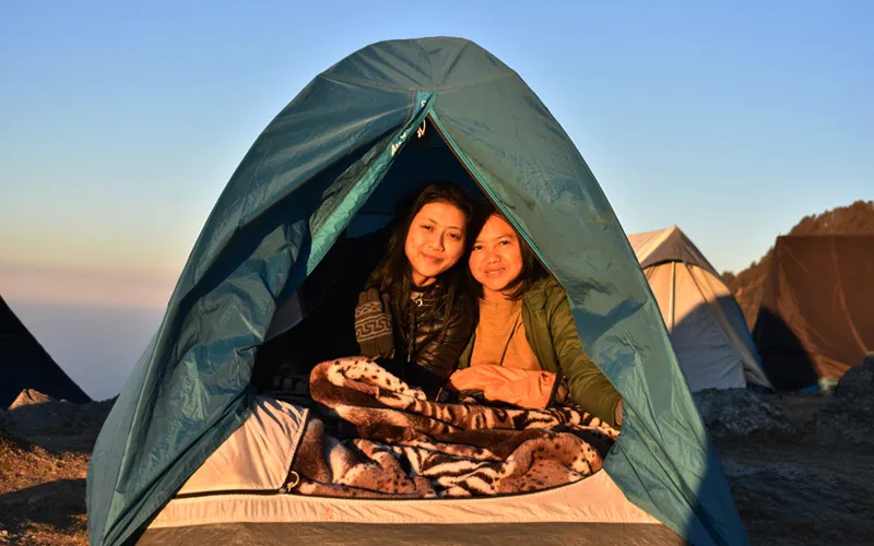 Two female campers smiling from inside a cozy camping tent pitched on the Triund ridge
