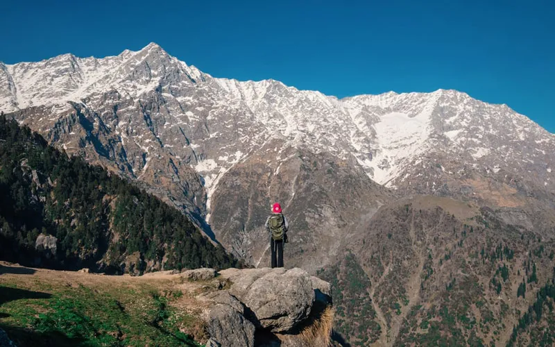 Trekker standing on a rocky ridge facing the snow-covered Dhauladhar mountain peaks in Triund